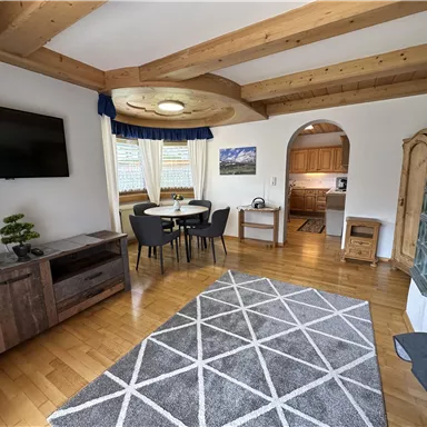 A cozy living space with a wooden beam ceiling and a large window. The room is furnished with a dining table, a sofa, and a carpet.