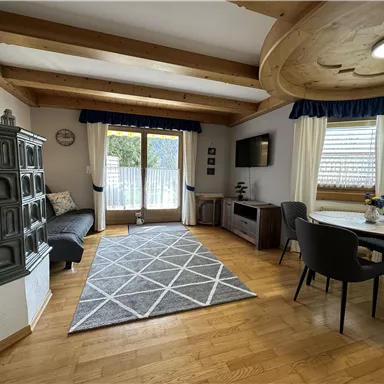 A cozy living room with a wooden beam ceiling and large windows. There is a couch, a table, and a modern television.
