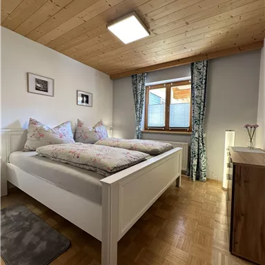 A cozy bedroom with a double bed and wooden ceiling. The room has a window with curtains and is bright and inviting.