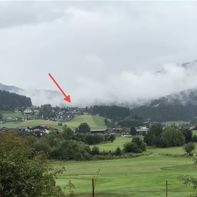 A picturesque landscape with gentle hills and a small village in the background. The sky is cloudy, lending a serene atmosphere to the scene.