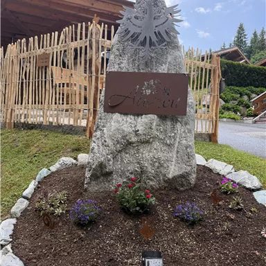 A large rock with a metal plaque displaying the name "Auen." Colorful flowers and stones decorate the area around the rock.
