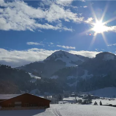 Eine schneebedeckte Landschaft mit Bergen im Hintergrund und strahlendem Sonnenschein. Ein rotes Gebäude steht im Vordergrund.