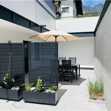 A modern terrace with a dining table and a sunshade.
Surrounded by decorative plants and a well-kept wall.