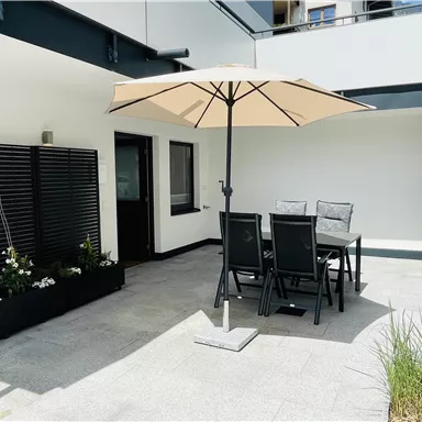 A modern terrace with a large sun umbrella and a dining area. In the background, plants and a simple wall can be seen.