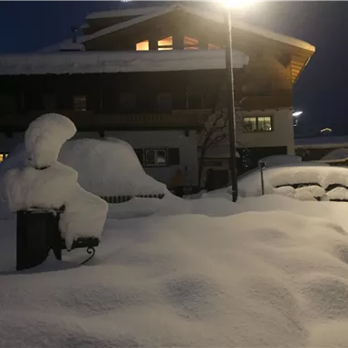Eine verschneite Landschaft bei Nacht mit einem beleuchteten Gebäude im Hintergrund. Der Schnee liegt hoch und bedeckt die Umgebung.