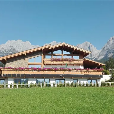 A picturesque house with a large balcony and colorful flowers. In the background, impressive mountains rise under a clear blue sky.