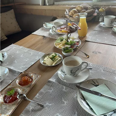 A beautifully set breakfast table with cups, dishes, jam, cheese, and fresh fruit. In the background, rolls and a glass of juice are visible.