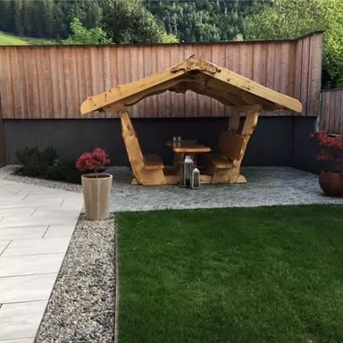 Ein einladender Garten mit einer Holzterrasse und einer großen Rasenfläche. Ein Tisch unter einem überdachten Sitzbereich lädt zum Entspannen ein.