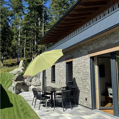 A terrace with a table and chairs, covered by a green sunshade. In the background, trees and a cozy house can be seen.