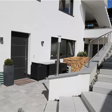 Ein modernes Haus mit einem klaren, weißen Außenanstrich und einer einladenden Eingangstür. Die Steintreppe führt zu einem gepflegten Außenbereich mit Holzlager und Pflanzen.