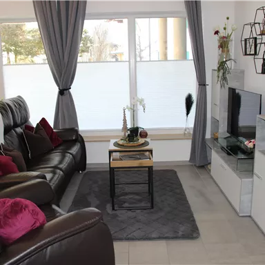 A modern living room with a comfortable black sofa and decorative cushions. The room has large windows and a television on a simple piece of furniture.