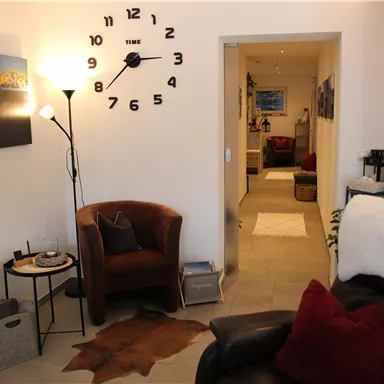 An inviting living room with a comfortable armchair, a table, and a large wall clock. The room has a bright, cheerful impression and leads to another area.