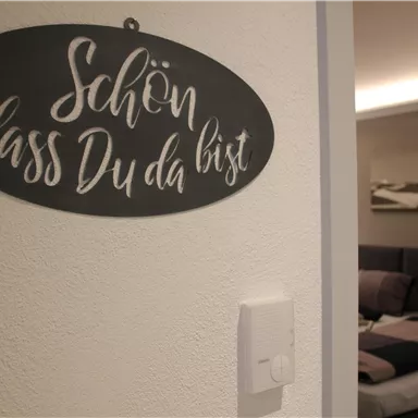 A welcome sign with the inscription “Nice to have you here.” In the background, there is a cozy room with a bed and soft colors visible.