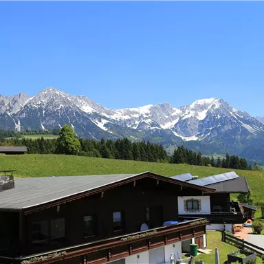 Eine wunderschöne Berglandschaft mit schneebedeckten Gipfeln und klarem, blauem Himmel. Vorne sind traditionelle Hütten in einer grünen Umgebung zu sehen.