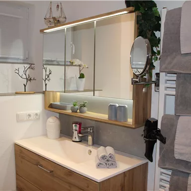 A modern bathroom with a wooden vanity and a large mirror. Decorative plants and bath accessories are placed on the shelf.
