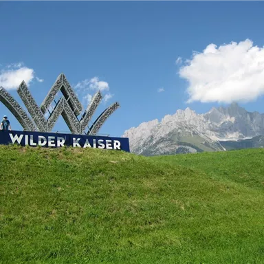Ein Schild mit der Aufschrift "Wilder Kaiser" steht auf einer grünen Wiese. Im Hintergrund sind majestätische Berge und ein blauer Himmel zu sehen.
