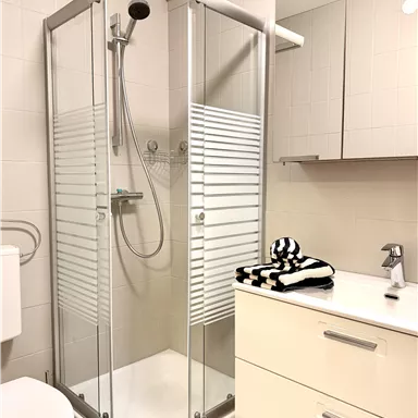 Ein modernes Badezimmer mit einer gläsernen Dusche und einem weißen Waschbecken. Auf dem Waschbecken liegt ein gestreifter Handtuch und die Wände sind hell gefliest.
