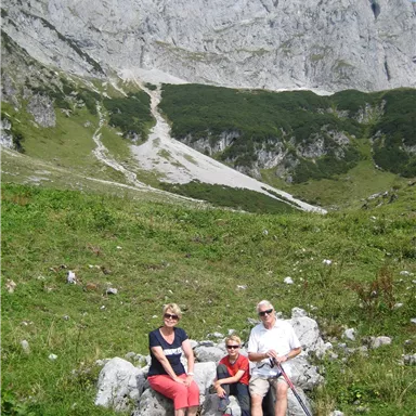 Eine Familie sitzt auf einem Felsen in der Natur. Im Hintergrund sind große Berge und eine grüne Wiese zu sehen.