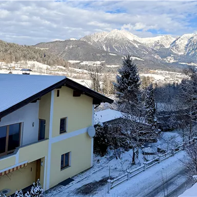 Ein charmantes Haus in einer verschneiten Landschaft. Im Hintergrund sind majestätische Berge und ein blauer Himmel zu sehen.