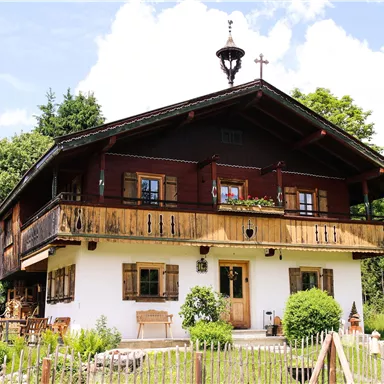 A charming old house with a wooden balcony and blooming windows. It stands in a green setting with trees and mountains in the background.