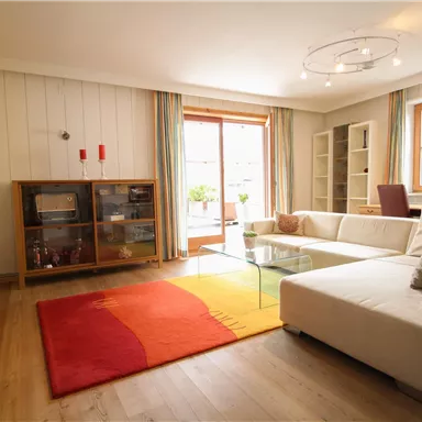 Ein modernes Wohnzimmer mit einem weißen Sofa und einem großen Glas Tisch. Der Raum hat große Fenster, die viel Tageslicht hereinlassen, und einen bunten Teppich auf dem Holzfußboden.