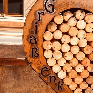 Ein rustikales Herz aus Holzscheiben mit der Aufschrift „Griaß enk“. Es befindet sich an einer Wand mit einem Fenster im Hintergrund.
