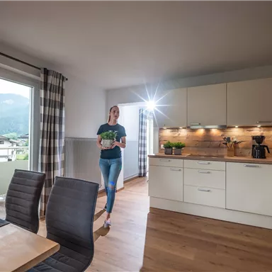 A modern kitchen with wooden and white furniture. A woman stands in the room holding a cup in her hand.