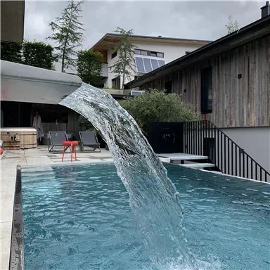 Ein moderner Pool mit klarem Wasser, aus dem ein Wasserfall fließt. Im Hintergrund sind Häuser mit Holzverkleidung sichtbar.