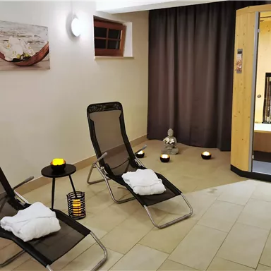 Ein entspannender Wellnessbereich mit zwei Liegen und sanfter Beleuchtung. Im Hintergrund ist eine Sauna zu sehen, die zur Ruhe und Erholung einlädt.