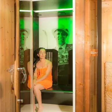 Eine Frau in einem orangefarbenen Handtuch sitzt in einer modernen Sauna mit grünem Licht. Im Hintergrund ist eine Darstellung einer Person zu sehen.