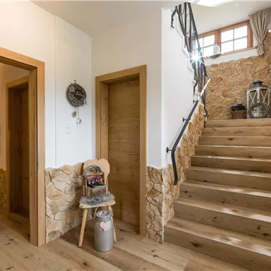 A bright corridor with wooden floors and stone walls. A staircase leads upstairs, and a cozy decoration is located in the corner.