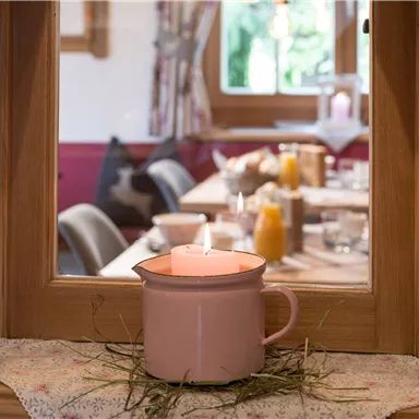 A cozy view through a window onto a laid table. A burning candle in a pink cup is in the foreground.