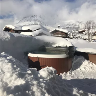 Ein verschneiter Garten mit einem Whirlpool und darunter liegender Schnee. Im Hintergrund sind Berge und ein blauer Himmel zu sehen.