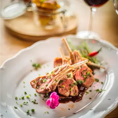 A delicious dish with lamb chops, garnished with a flower and herbs. In the background, a glass of red wine and a small bowl of side dishes can be seen.
