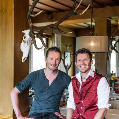 Two men are smiling while posing in a rustic restaurant with wooden decor and a deer antler in the background. They are wearing traditional attire and presenting an inviting atmosphere.