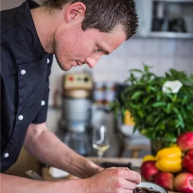 A chef is carefully preparing a dish in a modern kitchen. Fresh vegetables and kitchen utensils can be seen in the background.