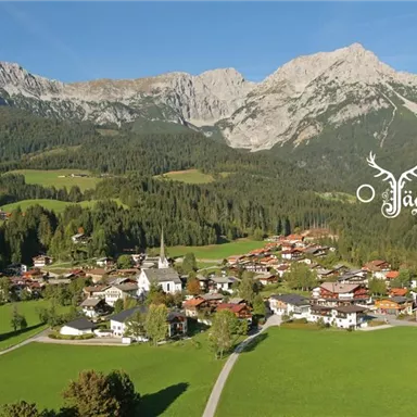 A picturesque landscape with a small village and high mountains in the background. Green meadows and forests surround the buildings.