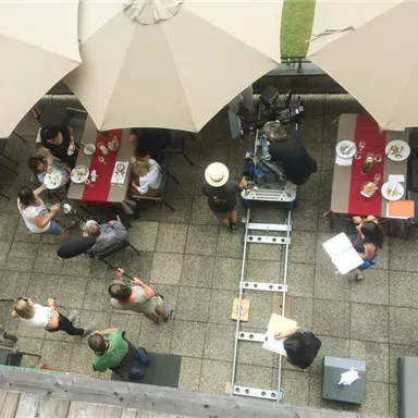 An outdoor terrace with several tables and parasols. Some guests are enjoying their meal while a film crew is taking footage.