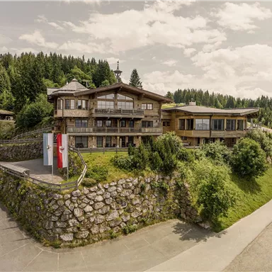 A modern wooden house surrounded by green landscape and trees. The road gently winds around the building.
