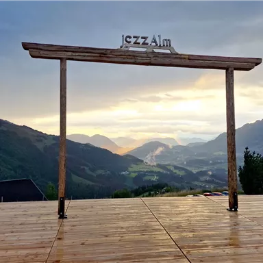 A scenic platform with a wooden beam on which "jazzAlm" is written. In the background, mountains and a beautiful sky at sunset can be seen.
