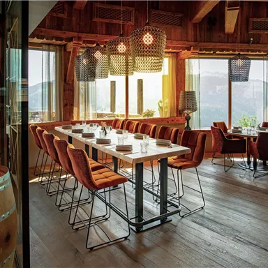 A stylish restaurant with wooden elements and large windows overlooking the mountains. The tables are elegantly set, and the chairs provide a comfortable seating option.