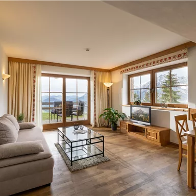 A cozy living room with a sofa, a table, and large windows. The room is bright and has a view of the nature outside.