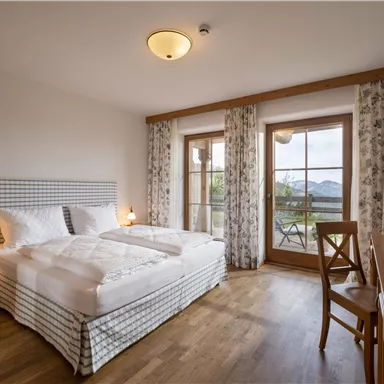 A modern bedroom with a double bed and wooden flooring. Large windows offer a view of nature.