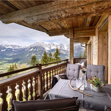A beautiful terrace with a wooden table and comfortable chairs. In the background, impressive mountains and green valleys extend.