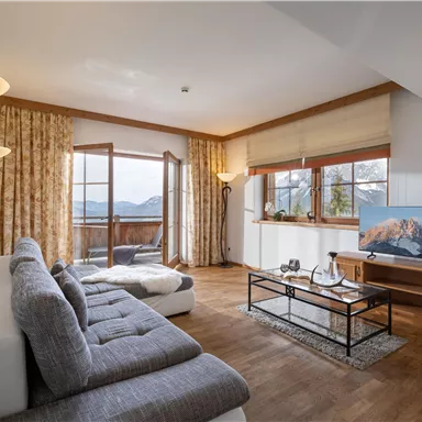 A modern living room with a cozy sofa and a television. Large windows offer a beautiful view of the mountains.