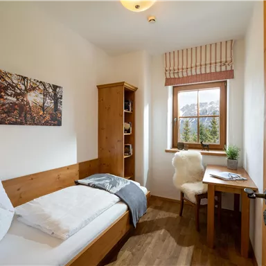 A cozy single room with wooden furniture and a window that offers a view of the mountains. The room is bright and friendly designed.