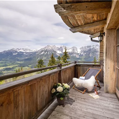 A cozy balcony with a view of the mountains and a green landscape. The area is equipped with wood and has a deck chair as well as decorated with plants.