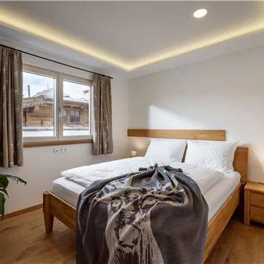 A cozy bedroom with a double bed and a modern wooden design. Windows let in natural light, and a plant creates an inviting atmosphere.