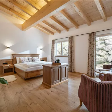 A bright bedroom with a wooden ceiling and a large bed. Two comfortable armchairs and windows with a view outside complete the room.