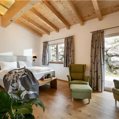 A modern bedroom with a wooden beam ceiling and large windows. There is a comfortable bed and a cozy armchair.